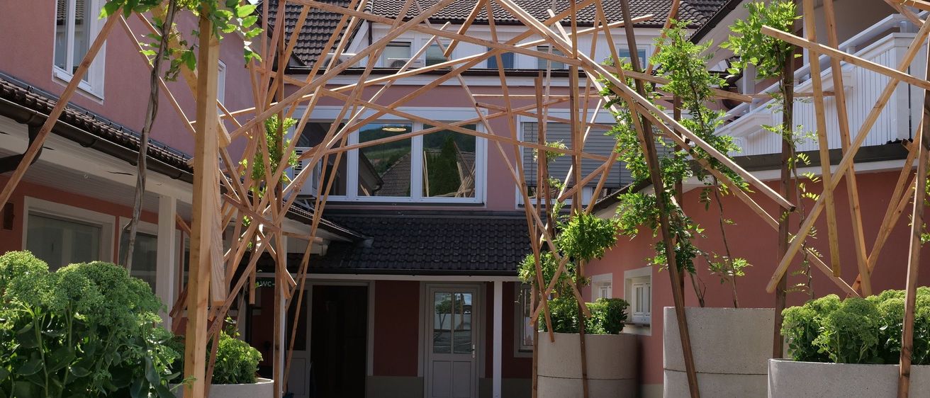 Eine Holzstruktur mit geometrischem Design steht vor einem Gebäude mit rosa Wänden und Fenstern. Es befindet sich in einem Innenhof mit Pflanzen in großen Betontöpfen.