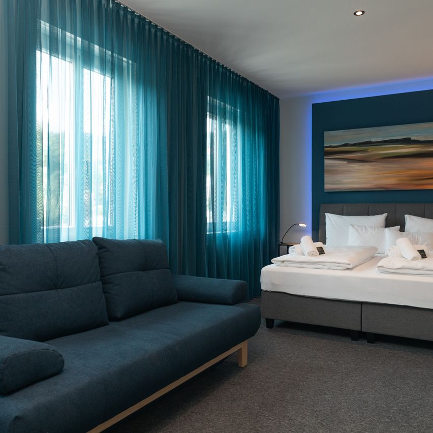 A blue-themed bedroom with a double bed and sofa. Blue curtains cover the windows, and a painting is on the wall.