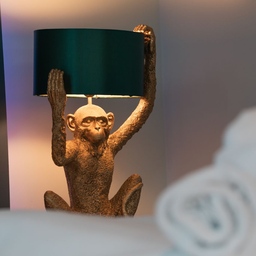 A golden monkey statue holds a green lampshade on a table, illuminated by a warm light. A white towel is placed to the right.