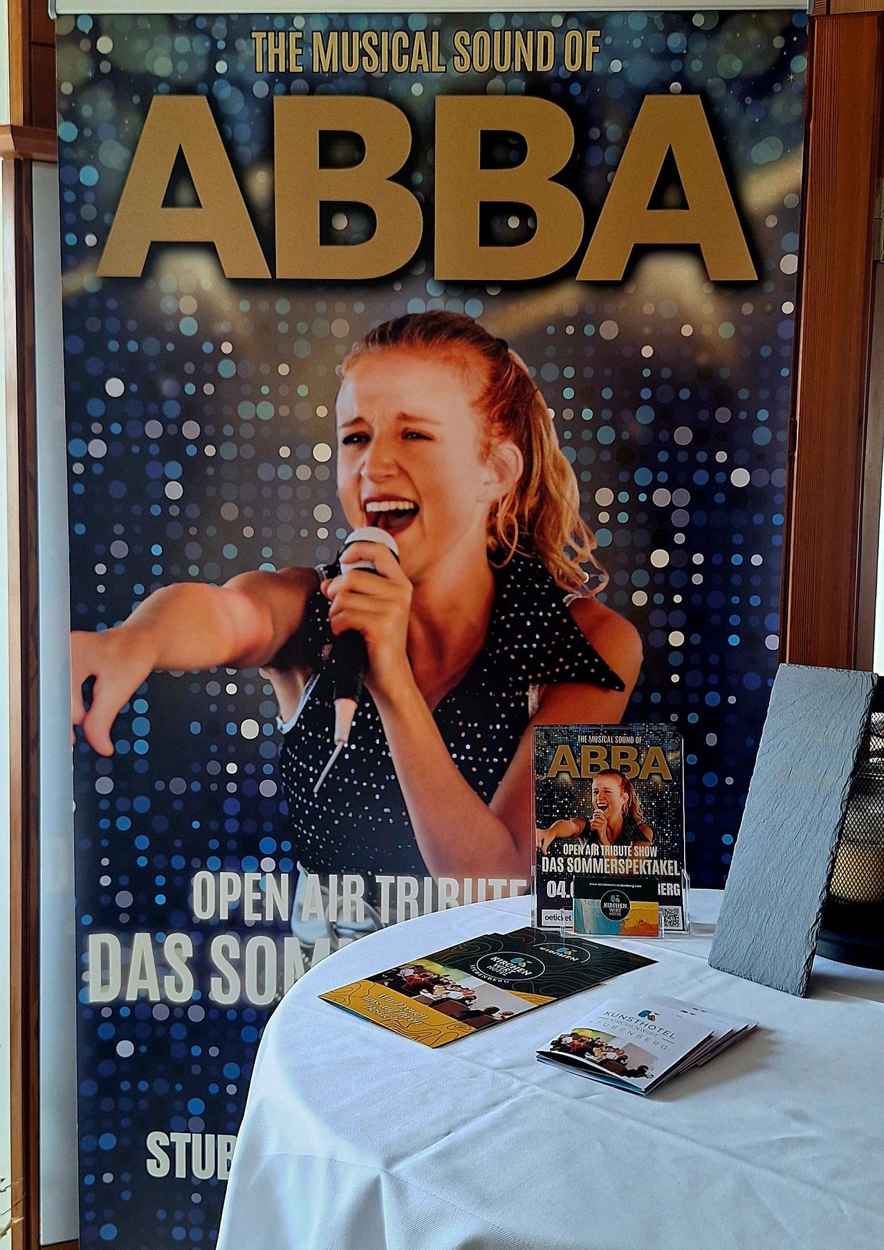 Ein Poster für eine Open-Air-ABBA-Tribute-Show zeigt eine singende Frau. Ein Tisch zeigt Broschüren und eine Zeitschrift, alles unter dem ABBA-Logo.