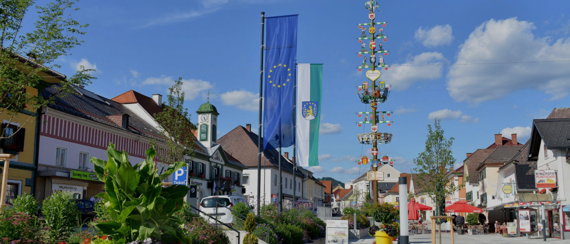 Ein Stadtplatz mit europäischen und deutschen Flaggen, bunten Blumen und einem dekorativen Pfahl. Im Hintergrund befinden sich Gebäude und ein Glockenturm.