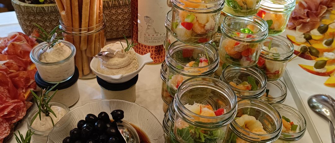 Ein Tisch mit Garnelensalat in Gläsern, Oliven, Brotsticks und einer Flasche Sarpa-Wein. Es gibt auch verschiedene andere Speisen, wie ein Fleischteller, und ein Tisch voller Garnierungen.