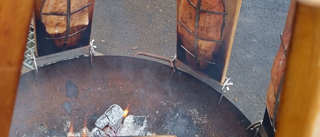Ein Outdoor-Kochsetup mit zwei Holzständern, die geräucherten Fisch halten, darunter lodernde Flammen.
