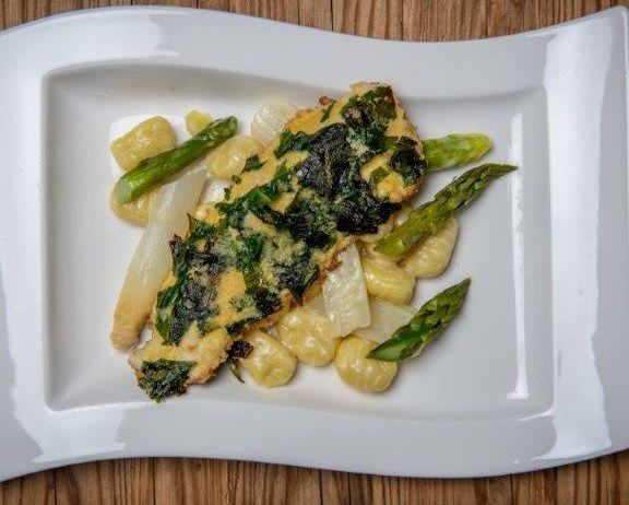 Ein weißer Teller mit einem Fischgericht, Spinat, Spargel und Pasta auf einer Holzoberfläche.