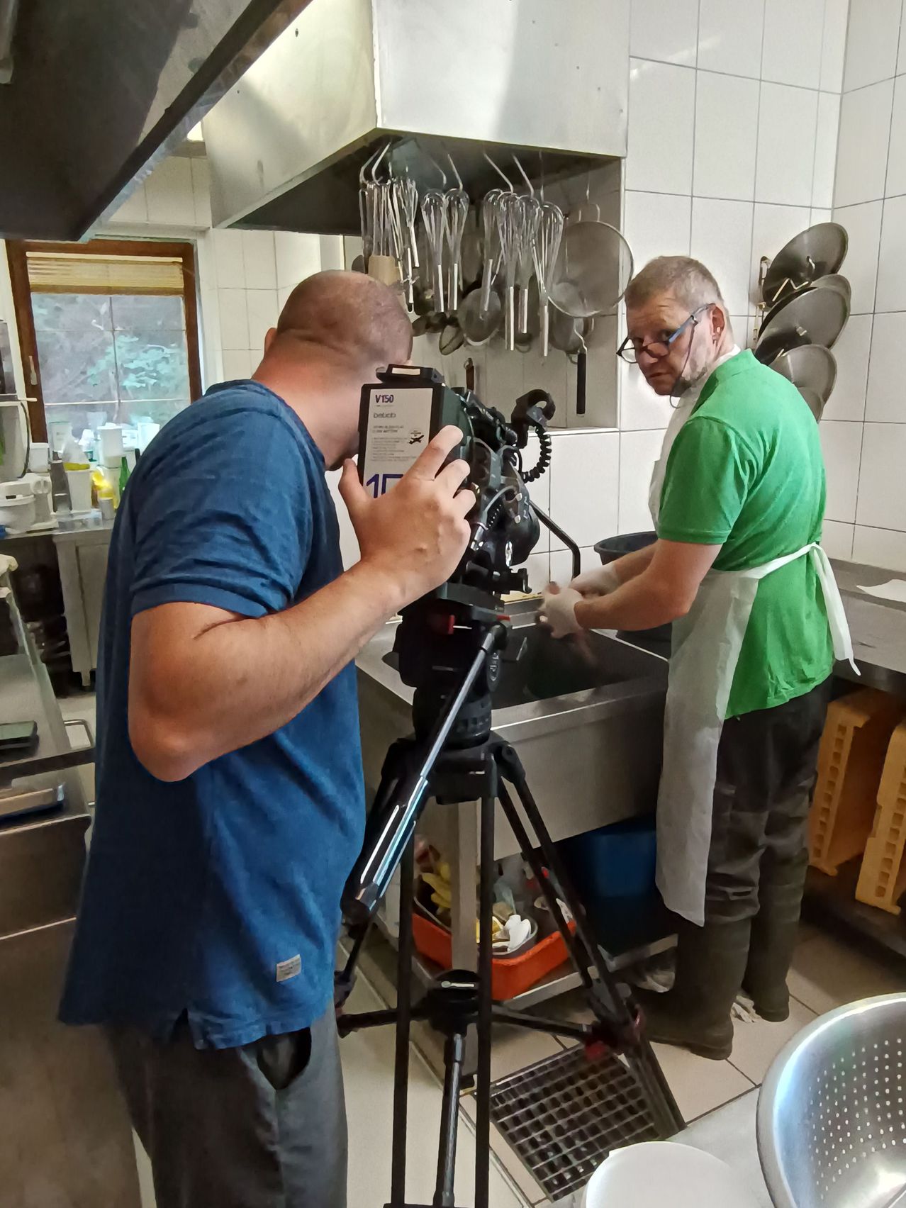 Ein Mann in einem blauen T-Shirt filmt einen anderen Mann in einem grünen T-Shirt, der in einer Küche Geschirr spült.