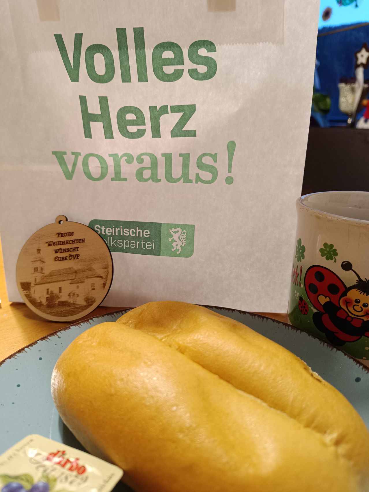 Ein Sandwich auf einem blauen Teller, mit einem Papierbeutel im Hintergrund, einer Holzmedaille und einer Kaffeetasse. Der Papierbeutel trägt den Aufdruck 'Voll Herz voraus!' und ein grünes Logo. Die Kaffeetasse hat ein Marienkäfer-Design.