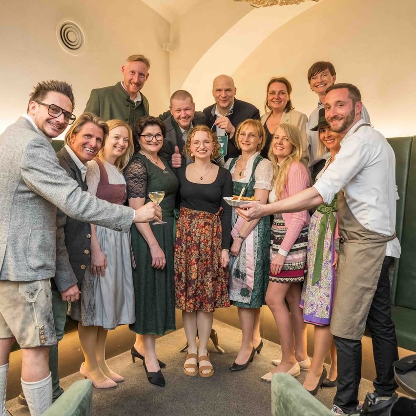 Eine Gruppe von Menschen in traditioneller Kleidung steht in einem Raum, lächelt und posiert für ein Foto. Einige halten Getränke und Teller mit Essen.