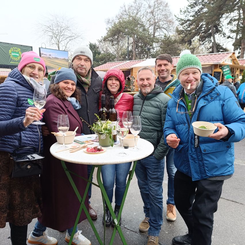 Eine Gruppe von Menschen steht um einen Tisch bei einer Outdoor-Veranstaltung, lächelt und hält Getränke. Sie tragen Winterkleidung.