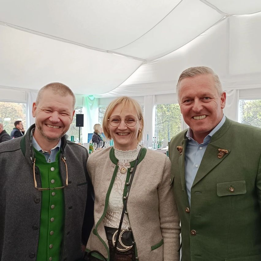 Drei Personen, zwei Männer und eine Frau, stehen gemeinsam in einem Zelt und lächeln für die Kamera. Der Mann auf der linken Seite trägt eine Brille und ein grünes Hemd. Die Frau in der Mitte trägt eine Brille und eine beige Jacke. Der Mann auf der rechten Seite trägt eine grüne Jacke. Hinter ihnen ist ein Lautsprecher sichtbar.