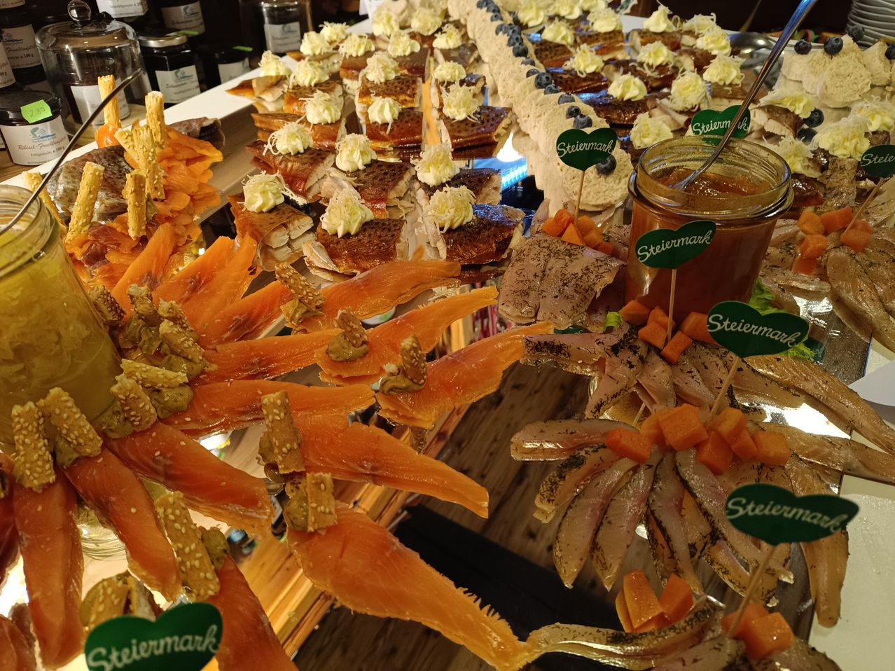 Eine Buffet-Tisch mit verschiedenen Gourmet-Gerichte, einschließlich Räucherlachs, Frischkäse und Blaubeeren. Der Tisch hat Holzoberflächen und ist mit grünen Etiketten dekoriert.