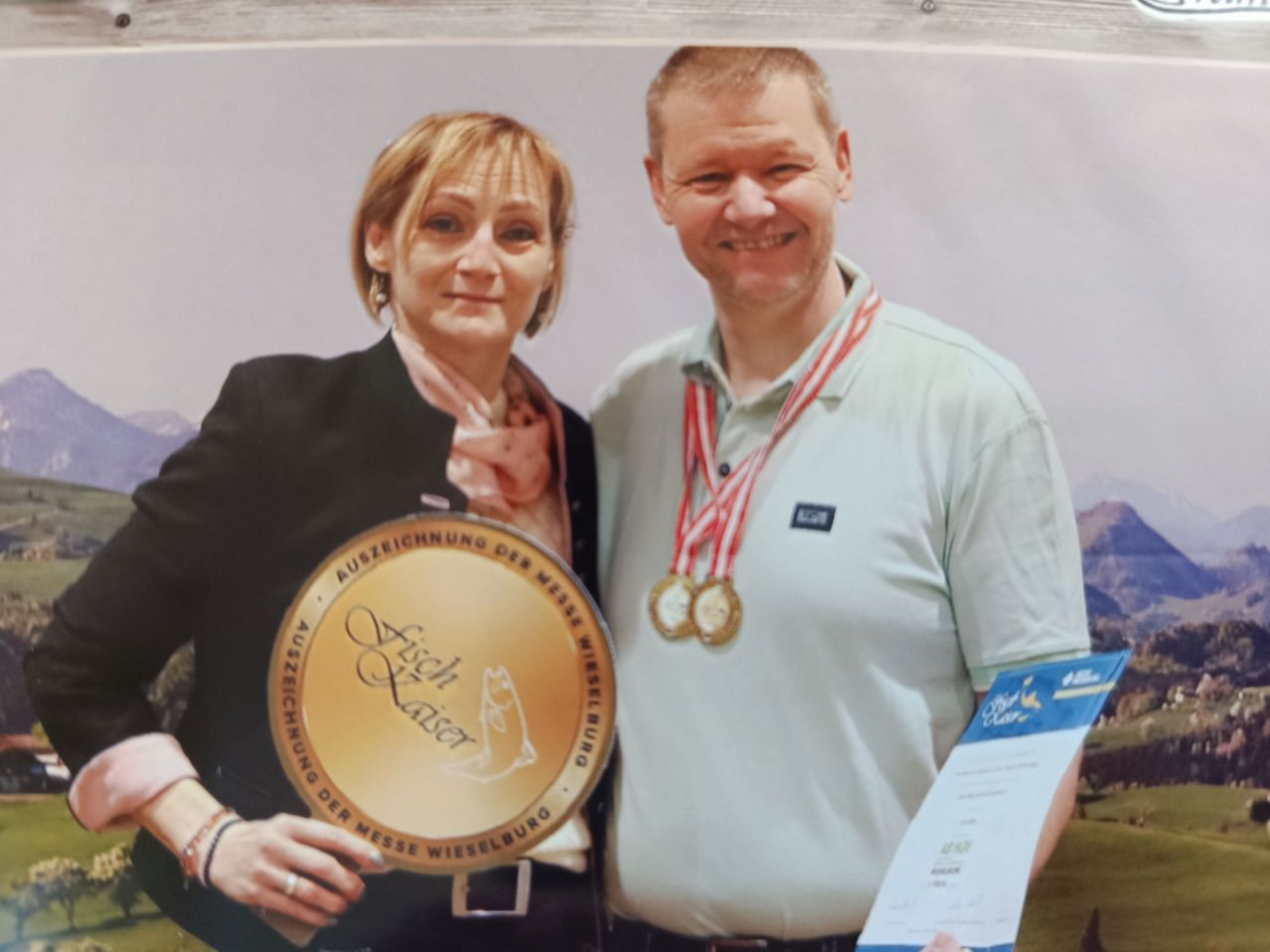 Ein Mann und eine Frau lächeln und posieren für ein Foto. Die Frau hält eine goldene Trophäe, und der Mann hat eine Medaille um den Hals. Sie scheinen beide glücklich und stolz zu sein.