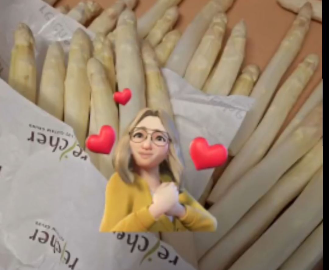 Ein Bild einer Frau mit Brille, die ihre Hände zusammenhält, umgeben von Bündeln Spargel mit roten Herzen im Hintergrund.