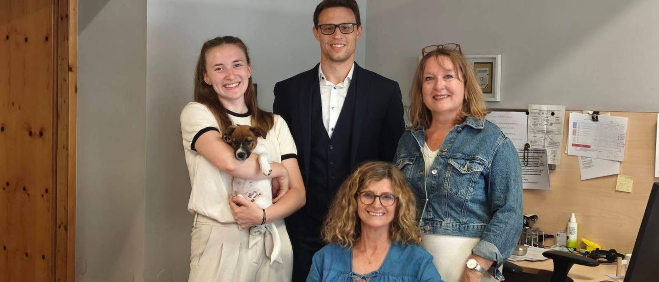 Fünf Personen posieren für ein Foto in einem Büro. Eine Frau hält einen Hund, und die anderen lächeln. Ein Laptop und Papiere liegen auf dem Tisch vor ihnen.