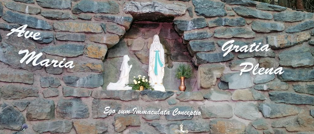 Ein steinerner Altar mit einer gewölbten Nische, die Statuen von Maria und Jesus zeigt, geschmückt mit Blumen, Kerzen und einem Kreuz. Lateinische Inschriften über der Nische lauten 'Gratia Plena' und darunter 'Ego sum Immaculata Conceptio'.