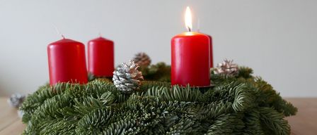Ein Weihnachtskranz aus Tannenzweigen mit drei roten Kerzen und einigen silbernen Tannenzapfen auf einer Holzoberfläche.