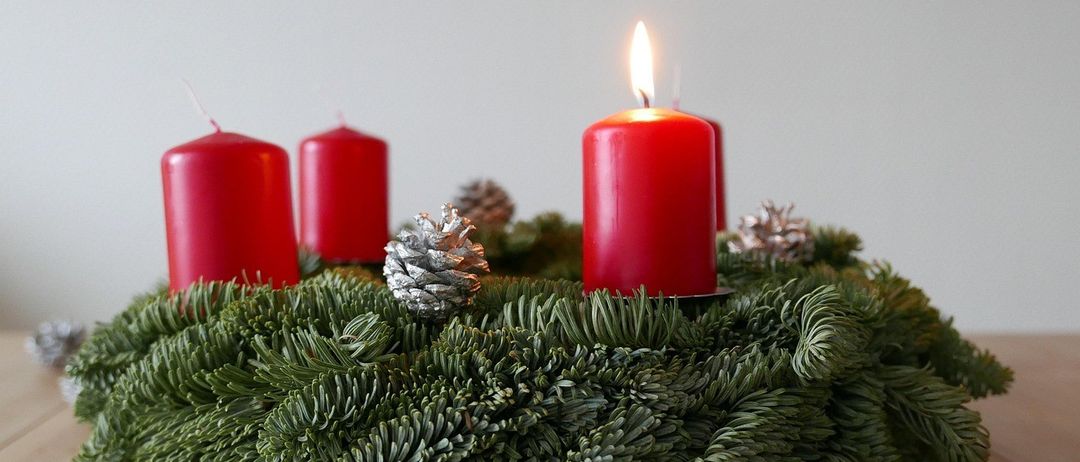 Ein Weihnachtskranz aus Tannenzweigen mit drei roten Kerzen und einigen silbernen Tannenzapfen auf einer Holzoberfläche.