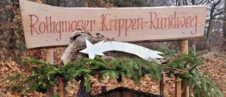 Ein Holzschild mit der Aufschrift 'Rothgnoser Krippen-Rundweg' über einer Weihnachtskrippe, umgeben von immergrünen Pflanzen und einem Stern.