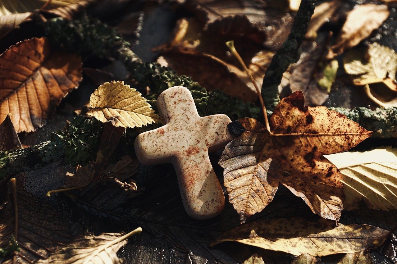 Ein kleines, verwittertes Kreuz liegt zwischen Herbstblättern, einige mit sichtbaren Flecken. Das Kreuz besteht aus Stein und ist teilweise mit Schmutz bedeckt.