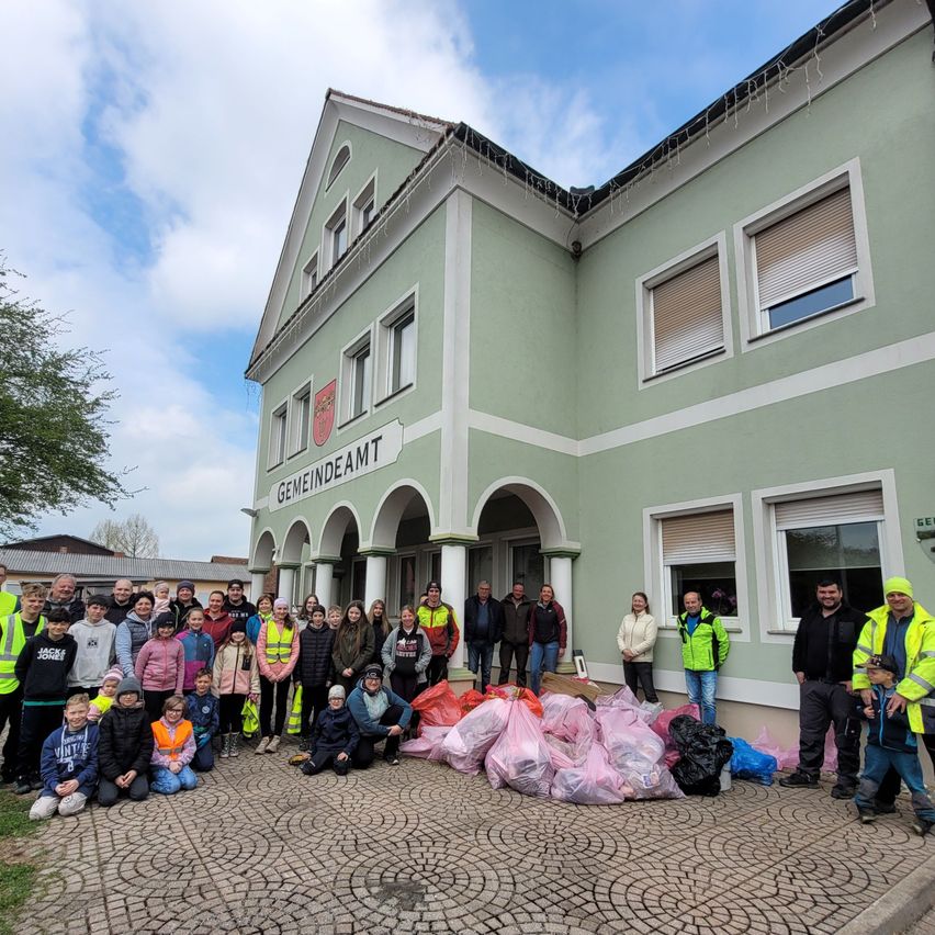 A group of people in safety vests pose for a photo in front of a building with the name Gemeindeamt. The building has several windows and a red logo on the wall. Several bags of garbage are in front of them.