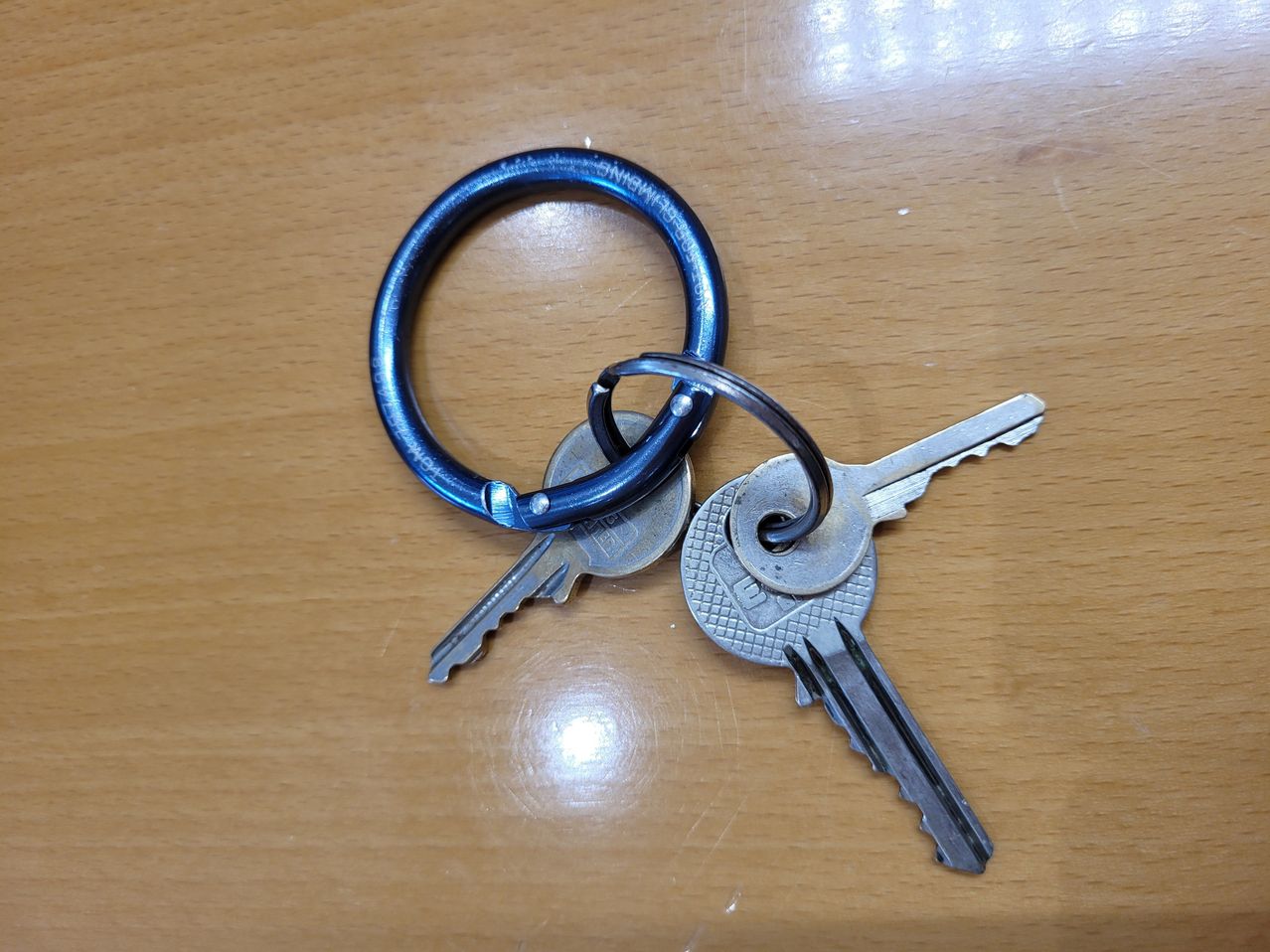 Ein Paar silberner Schlüssel mit einem blauen Schlüsselring auf einer Holzoberfläche. Die Schlüssel haben eine flache Oberfläche mit Rillen und einem einzigartigen Design.
