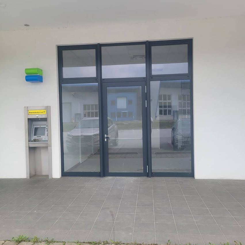 Eine Außenansicht eines modernen Gebäudeeingangs mit doppelten Glastüren, einem Geldautomaten und einem in der Scheibe reflektierten Auto.