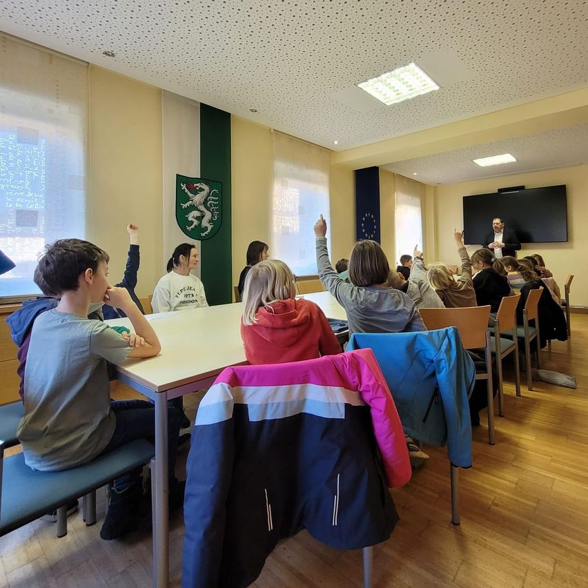 Ein Klassenzimmer mit Holzboden und grüner Wand hat Schüler, die an Tischen sitzen, mit Jacken, die auf den Stuhlrücken hängen. Ein Mann hält vorne im Raum eine Präsentation.
