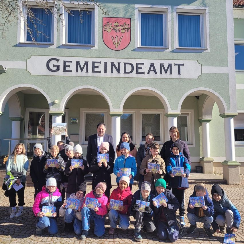 Eine Gruppe von Kindern und Erwachsenen posiert für ein Foto vor einem Gebäude mit dem Schild Gemeindeamt. Sie halten alle bunte Broschüren.