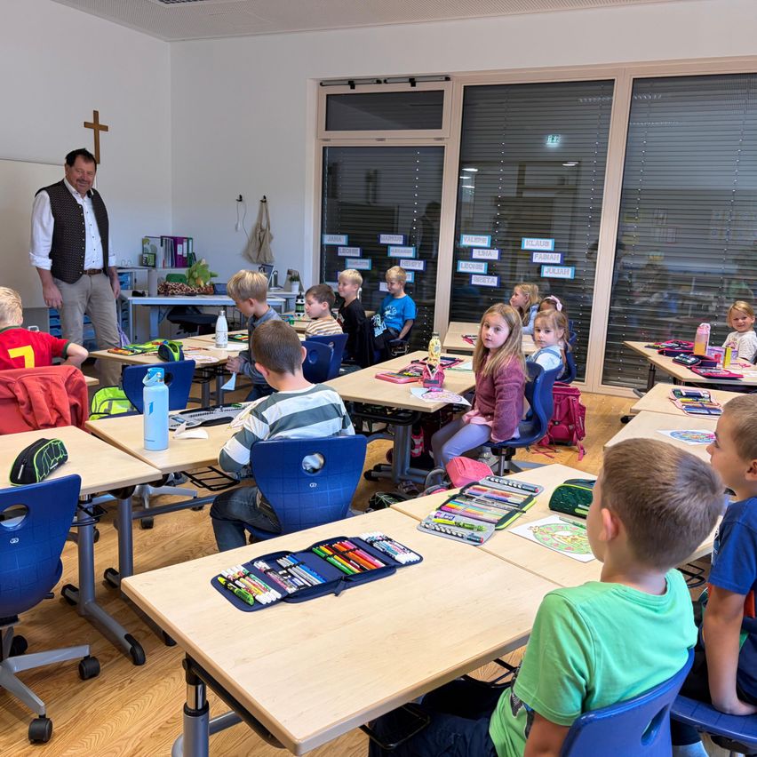 Ein Klassenzimmer mit Kindern an Schreibtischen, ein Lehrer steht vorne und es gibt verschiedene Schulsachen auf den Schreibtischen.
