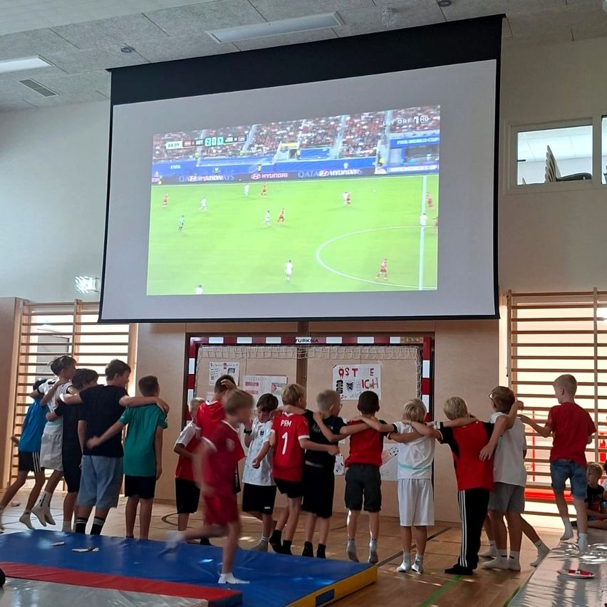 A group of children in sports jerseys gather around a large screen displaying a soccer match. They stand on a blue mat, possibly inside a gym.