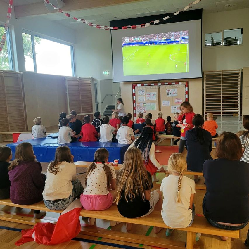 Children are seated on benches watching a soccer game on a projector screen. Some have snacks on small plates.
