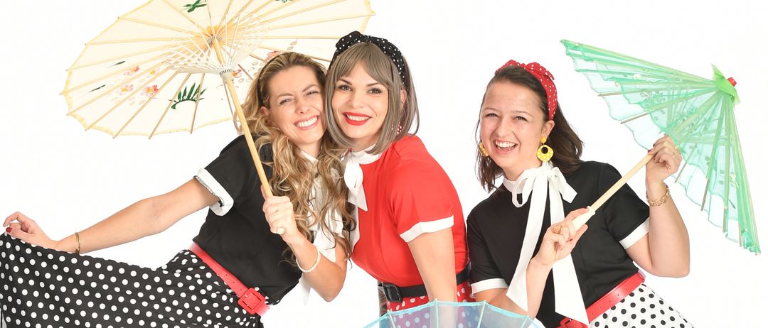 Drei Frauen in Retro-Kleidung stehen eng beieinander und lächeln. Jede hält einen Schirm, einer mit einem Blumendesign. Sie tragen gepunktete Röcke, Gürtel und schwarze Stiefel.