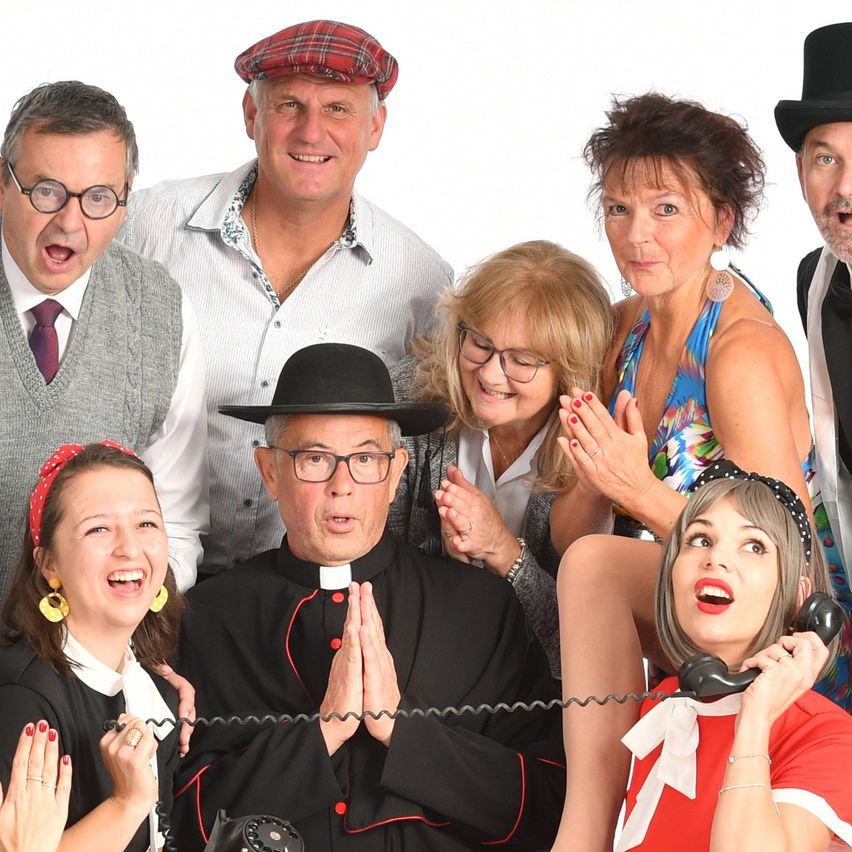Eine Gruppe von Erwachsenen in verschiedenen Kostümen, einschließlich eines Priesters, posiert für ein Foto. Einige tragen Hüte, Brillen und Schmuck und sie alle lächeln und machen lustige Gesichter.
