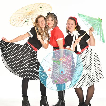 Drei Frauen in Retro-Kleidung, lächelnd, posieren mit Regenschirmen. Jede Frau trägt einen Punkterock und ein Hemd, eines in Schwarz, eines in Rot und eines in Blau. Sie halten bunte Regenschirme mit Blumenmustern.