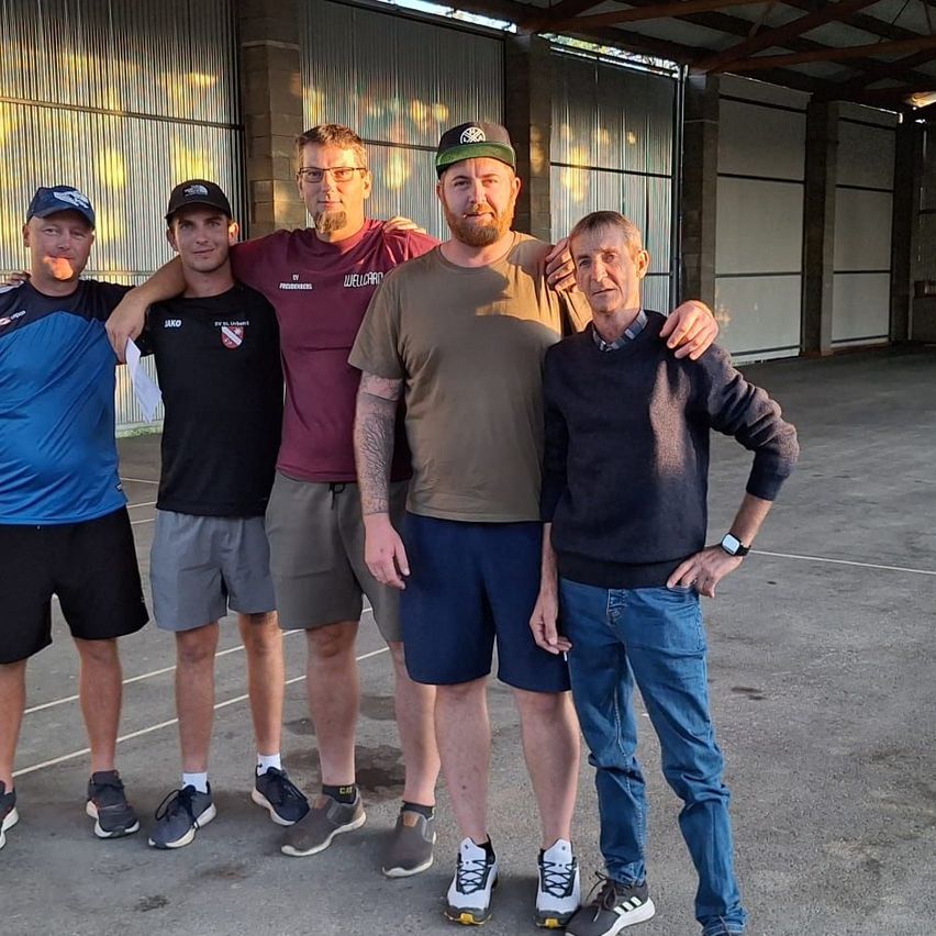 Fünf Männer stehen in einer Garage und lächeln für ein Foto. Einer trägt ein burgunderrotes Shirt, ein anderer eine grüne Kappe. Sie sind lässig gekleidet, mit Turnschuhen und Shorts.