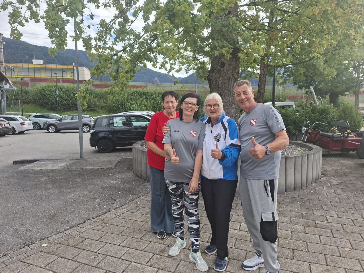 Vier Personen, zwei Frauen und zwei Männer, stehen zusammen auf einem Parkplatz, lächeln und zeigen Daumen hoch. Sie tragen Freizeitkleidung und stehen vor einem Baum und einem Betonpflanzer. Hinter ihnen sind geparkte Autos, ein Gebäude und eine Bergkette.