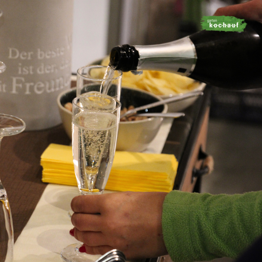 Eine Flasche Champagner wird in eine Sektflöte auf einem Tisch mit Snacks und gelben Servietten gegossen. Ein Keramikkrug mit dem Text 'Der beste ist der Freund' ist im Hintergrund.