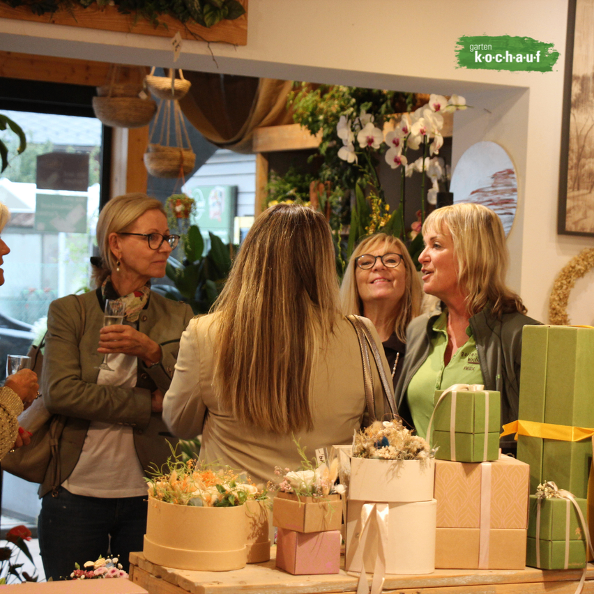 Vier Frauen stehen in einem Raum, lächeln und plaudern. Ein Tisch mit Blumen und verpackten Geschenken steht vor ihnen. Der Raum hat Pflanzen und ein Schild mit der Aufschrift 'garten ko-chauf'.