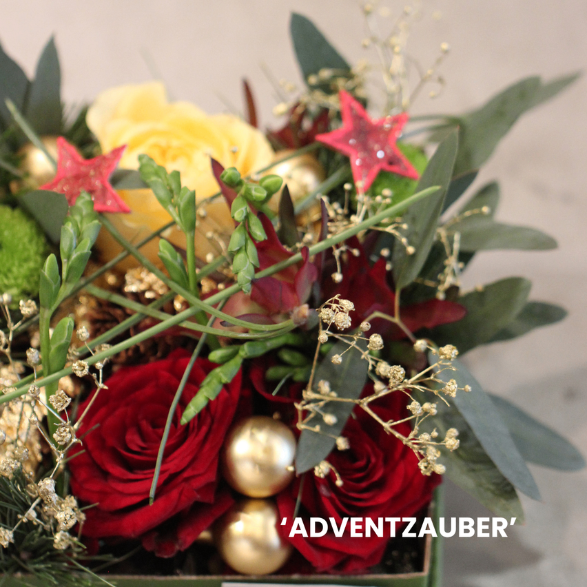 Ein Blumenarrangement mit roten Rosen, einer gelben Rose, goldenen Ornamenten und roten Sternen. Das Arrangement ist mit 'ADVENTZAUBER' beschriftet.