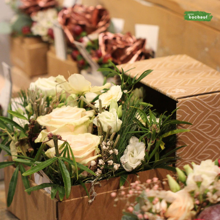 Eine braune Blumenarrangement-Box zeigt weiße Rosen und Grünpflanzen, platziert in einer Umgebung mit anderen ähnlichen Boxen und Kupferakzenten.