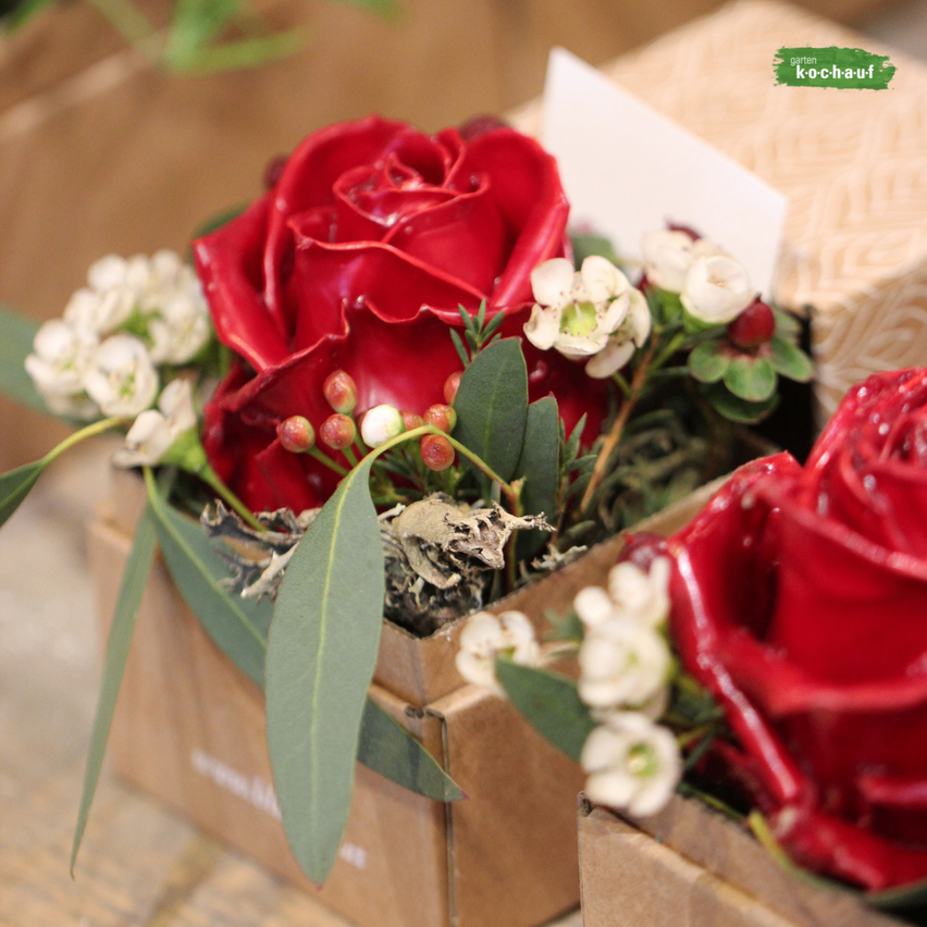 Zwei rote Rosen mit weißen Blumen und grünen Blättern in einer braunen Box auf einer hölzernen Oberfläche.