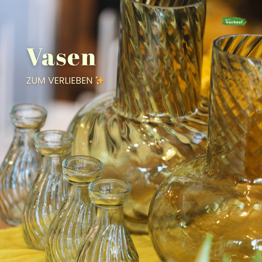 Eine Sammlung goldenfarbener Glasvasen, elegant auf einem gelben Tuch angeordnet. Der Text 'Vasen zum Verlieben' deutet an, dass die Vasen zum Verlieben gedacht sind.