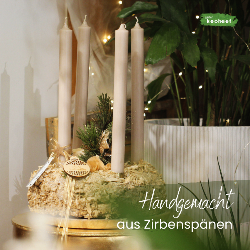 A handcrafted arrangement with four tall candles placed on a mossy base, adorned with greenery and a golden ribbon, in front of a plant with fairy lights.