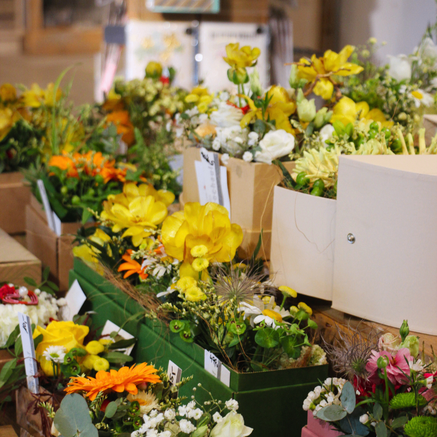Eine Vielzahl von Blumen ist in Schachteln mit Etiketten angeordnet. Die Blumen sind in verschiedenen Farben, einschließlich Gelb, Orange und Weiß. Es gibt auch einige grüne Blätter in der Anordnung.