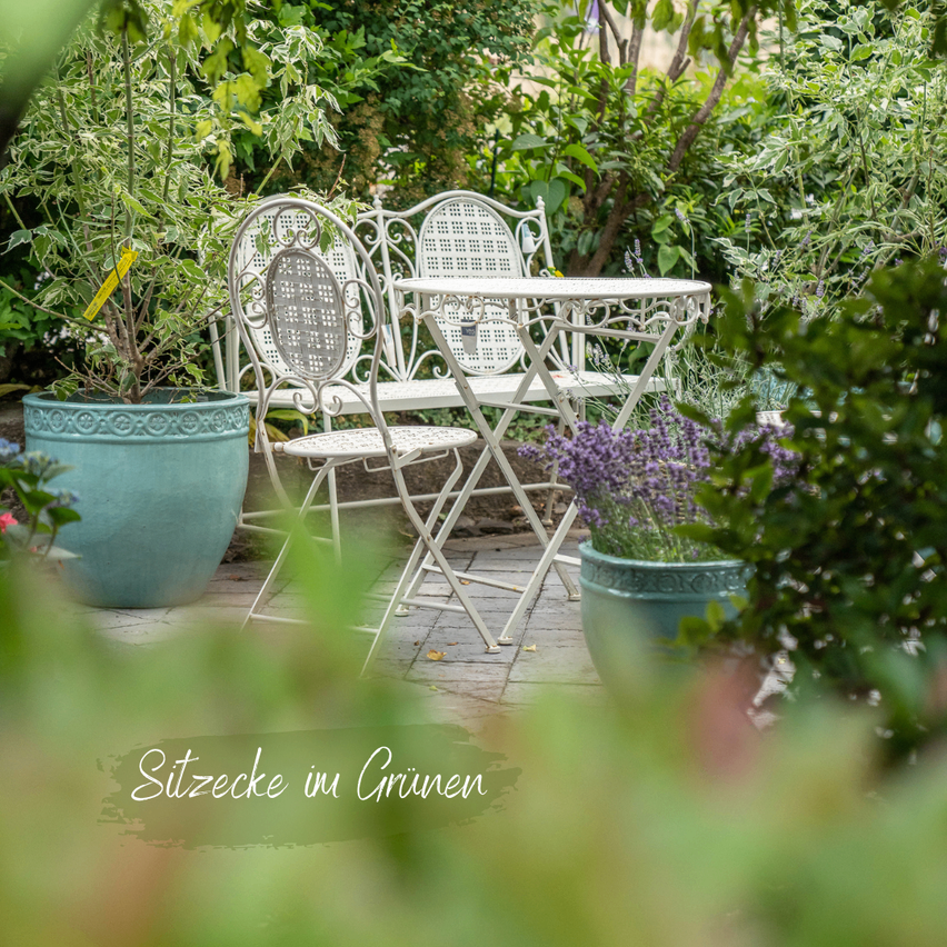 A garden setting with white metal chairs and a table surrounded by lush green plants and flowers. The image has a soft focus effect and a text overlay reading 'Sitzecke im Grünen'.