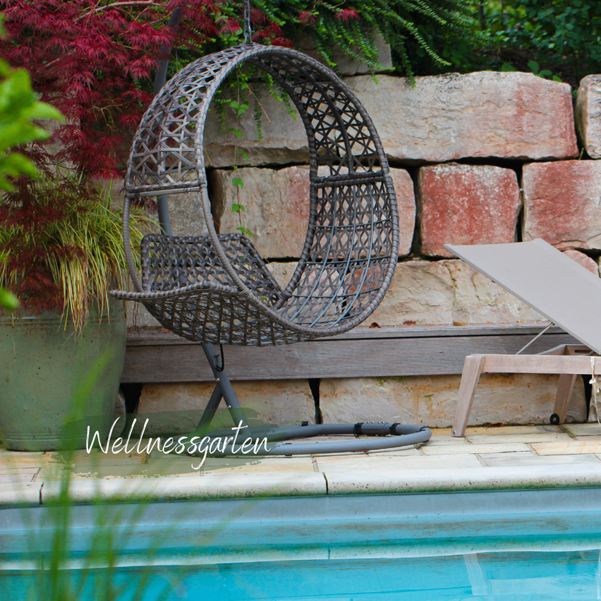 A round wicker chair hangs from a tree near a pool, with a lounge chair and a stone wall in the background.