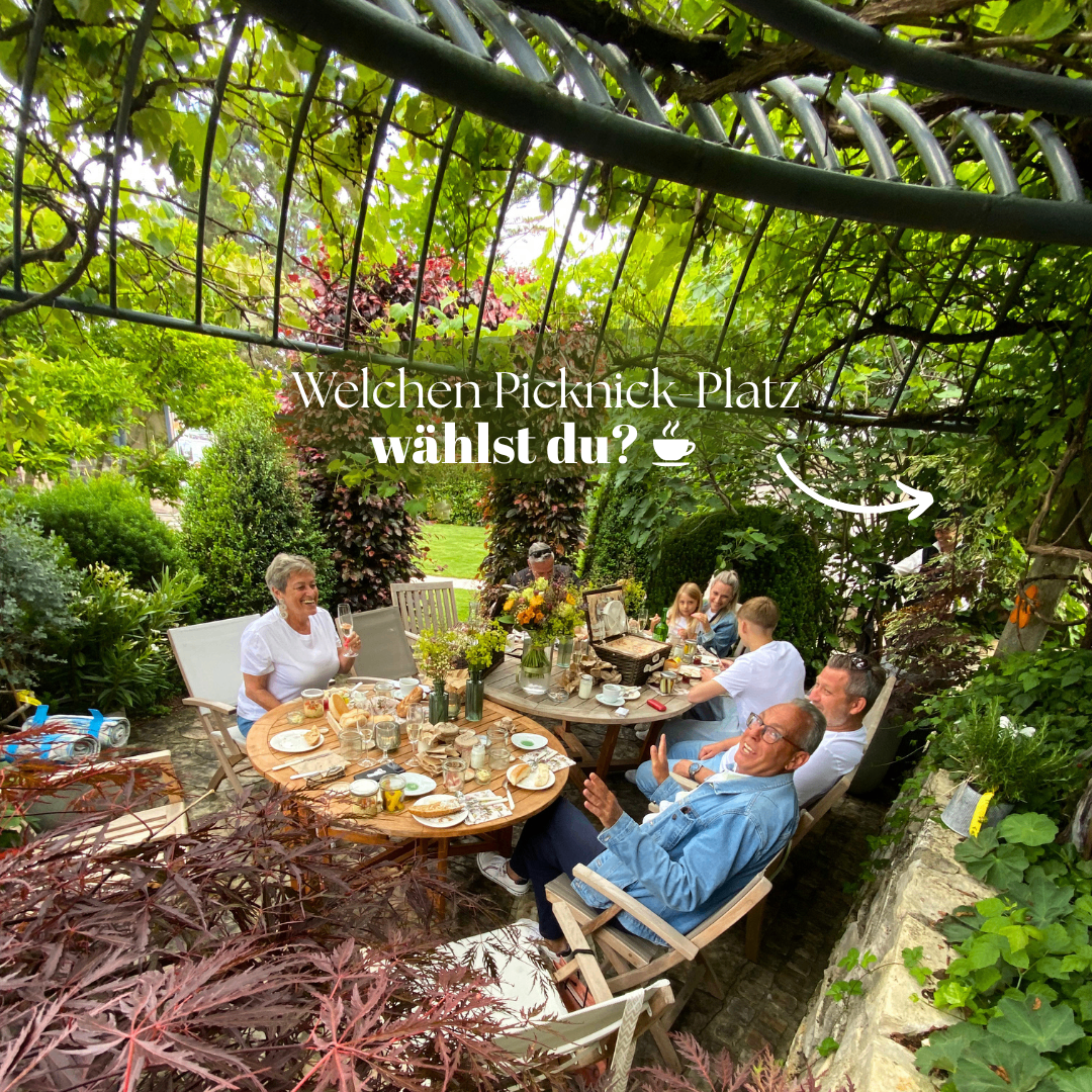 A family is enjoying a picnic in a garden with a table set for a meal, surrounded by greenery and a metal arch. The text 'Welchen Picknick Platz wählst du?' is displayed above.