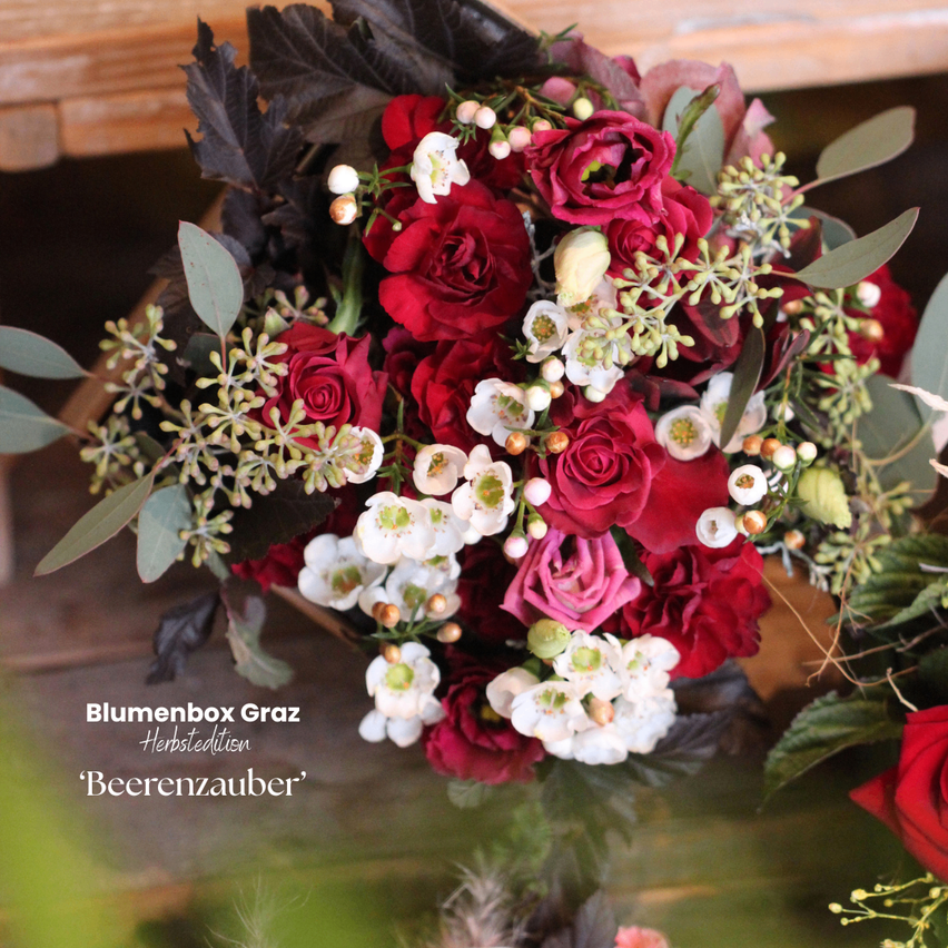 Ein Nahaufnahme eines lebhaften Blumenstraußes mit tiefroten Rosen, weißen Blüten und dunklen grünen Blättern. Der Strauß ist kunstvoll mit einer Mischung aus Texturen und Farben arrangiert.