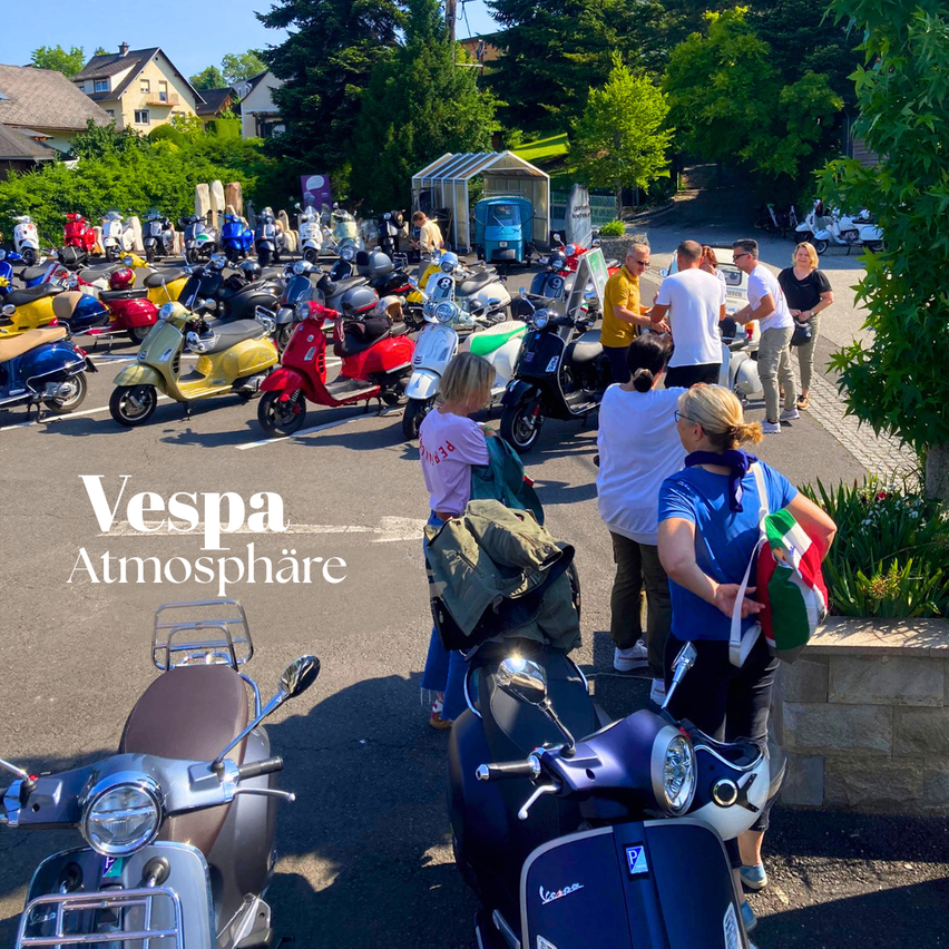 Various Vespa scooters parked in a parking lot with a building in the background. People are standing and conversing near the scooters.