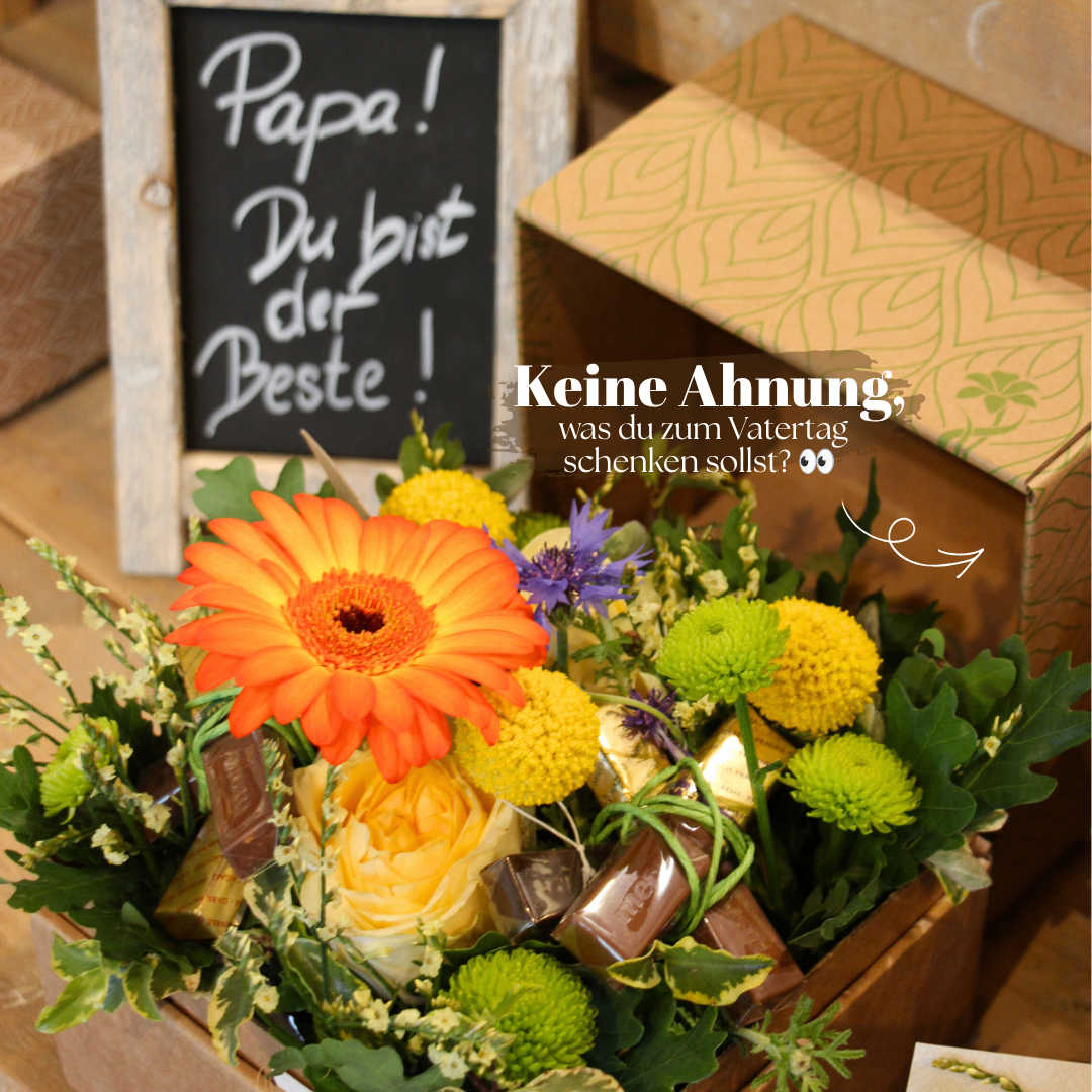 A flower arrangement with chocolates in a wooden box, accompanied by a chalkboard with 'Papa! Du bist der Beste!' and a decorative box with 'Keine Ahnung, was du zum Vatertag schenken sollst?'.