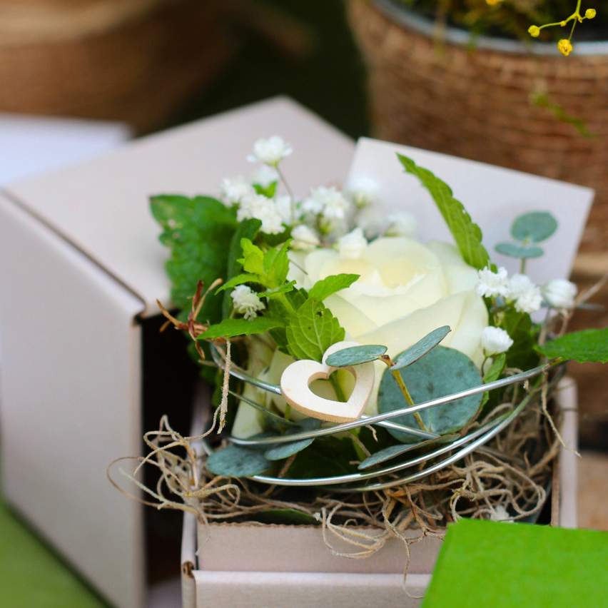 A white rose, surrounded by greenery, lies in a white box with a heart-shaped wooden decoration. The box is partially open.